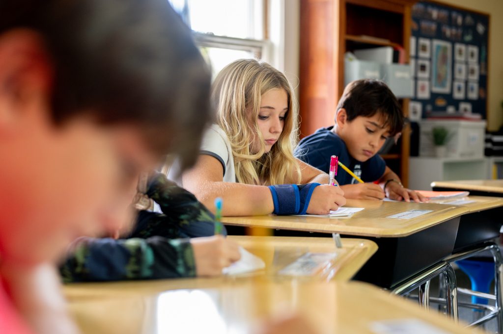 Middle school students working at desks with paper and pencil.