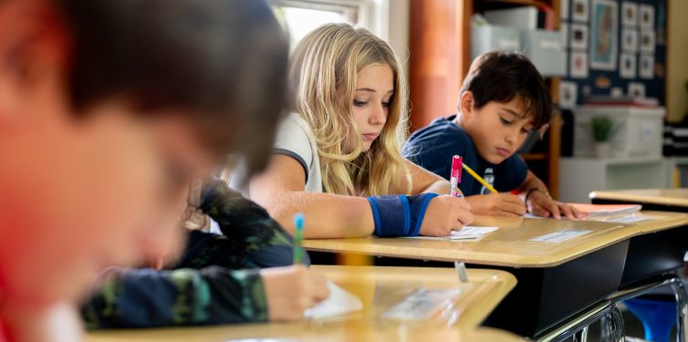 Middle school students working at desks with paper and pencil.