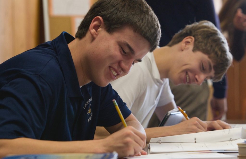 A smiling high school student takes notes at a desk.