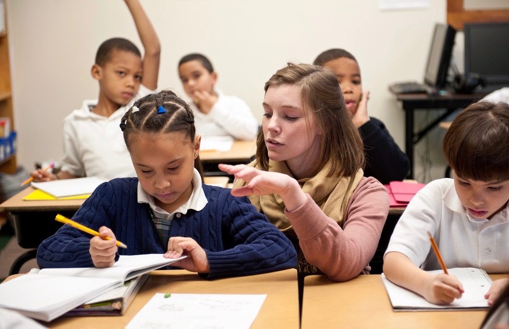 A teacher kneeling beside a desk to help an elementary student while other students in the back row of desks have their hands raised.