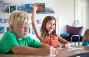Two students in an elementary classroom with a smiling student raising her hand.