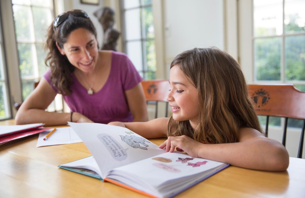 An elementary aged student is reading from a picture book alongside a smiling educator.