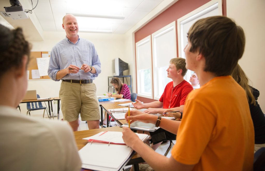 A standing high school teacher speaks to a classroom of students in a horseshoe arrangement of desks.