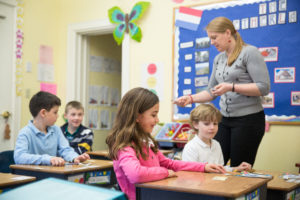 A teacher is handing out manipulative for a hands on activity to student seated in rows within her elementary classroom.