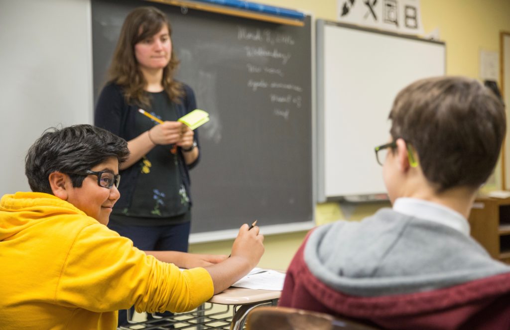 A teacher stands in front of a chalk board and speaks with middle school studen.ts