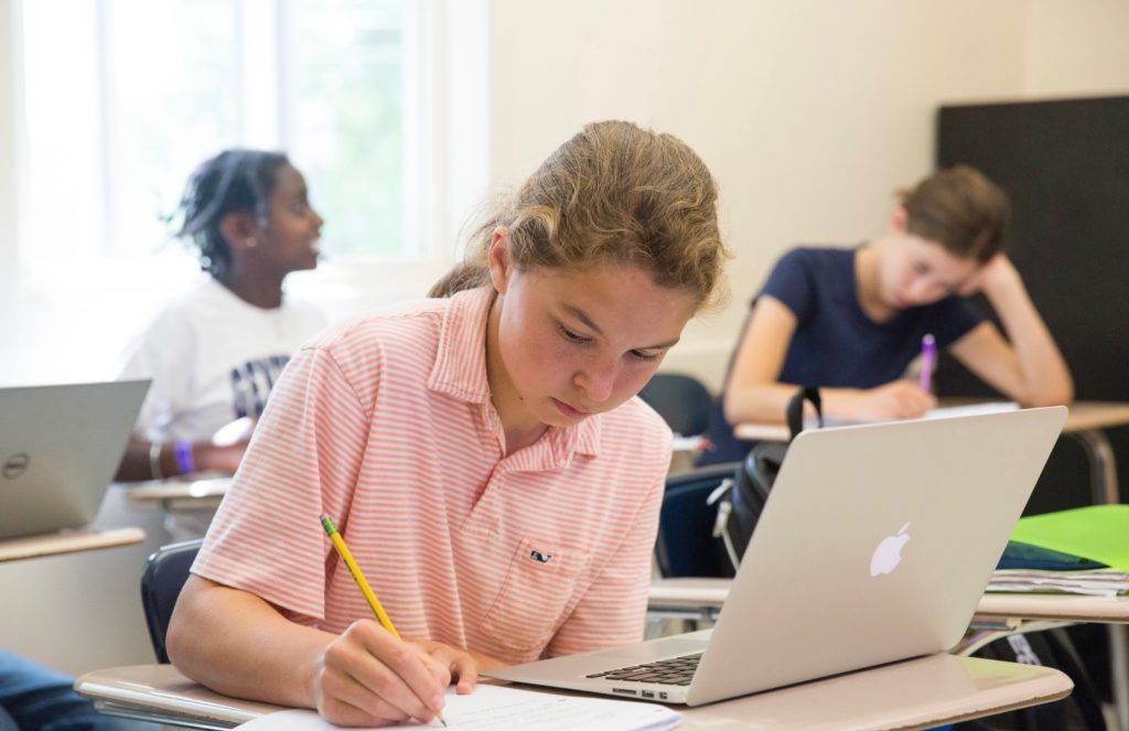 A high school student writes at a desk with an open computer.