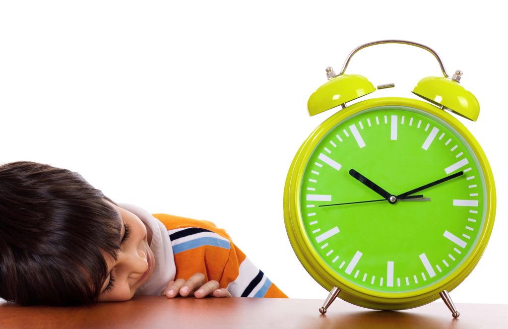 A young child asleep on a desk next to an analog clock.