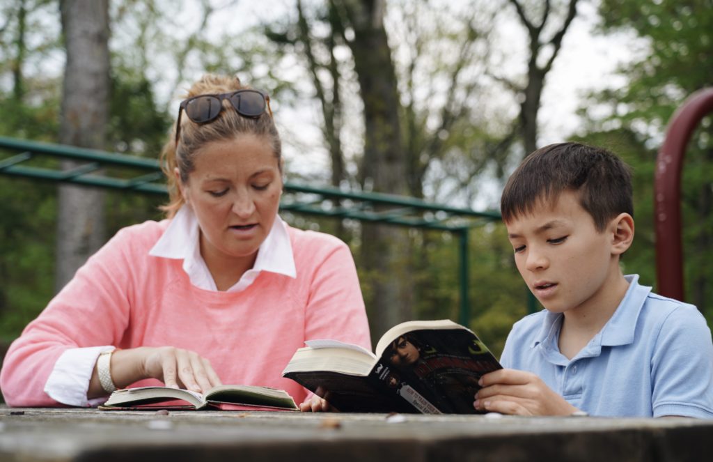 A teacher and elementary student are seated outside reading orally from copies of the same book.