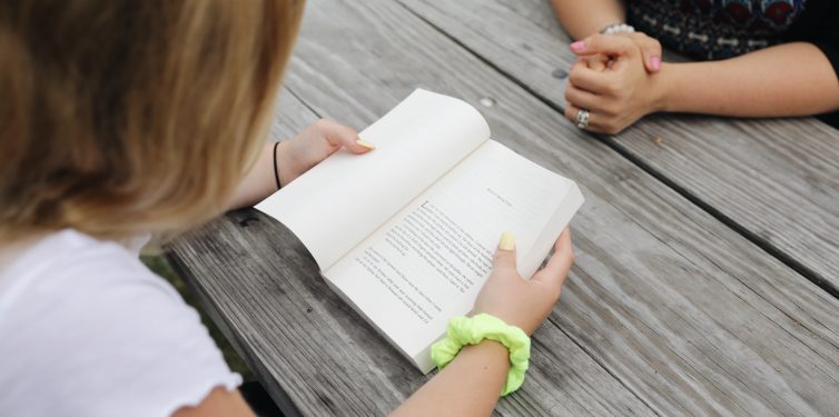 Bird's eye view of student reading a book sitting at a picnic table with adult sitting across from them.