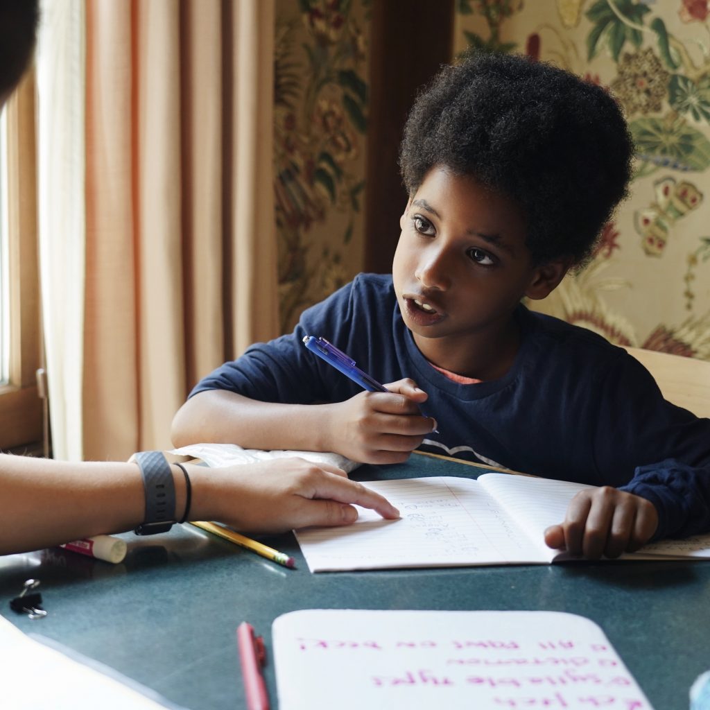 A teacher and an elementary age student are seated side by side. The student is writing while the teacher points out something on the page.