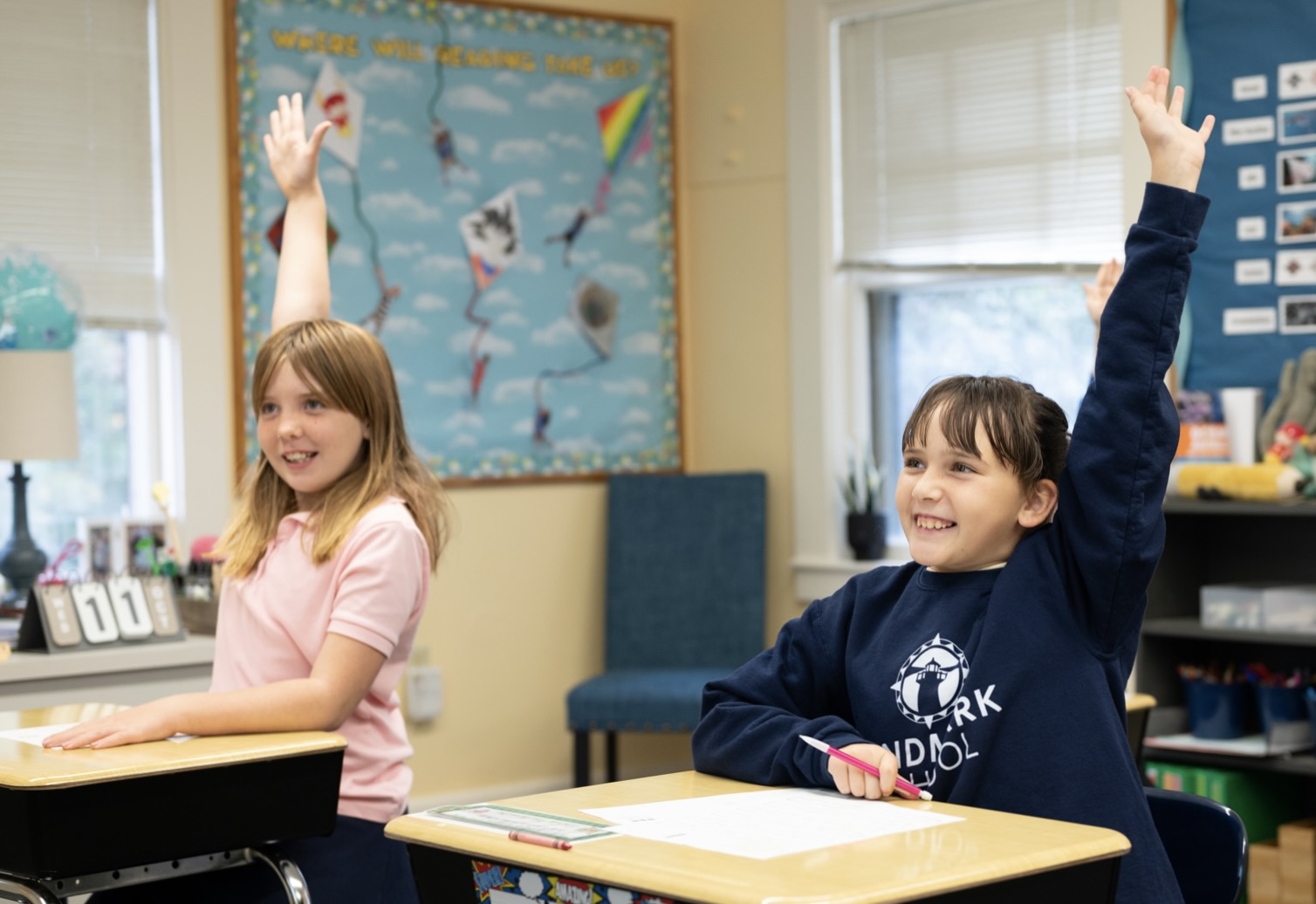 Two girls in a classroom raising their hands.