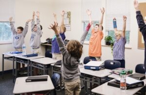 Students in an elementary classroom are all standing behind their desks with both arms reaching above their heads.