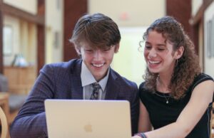 Two seated high school students smiling in front of an open laptop.