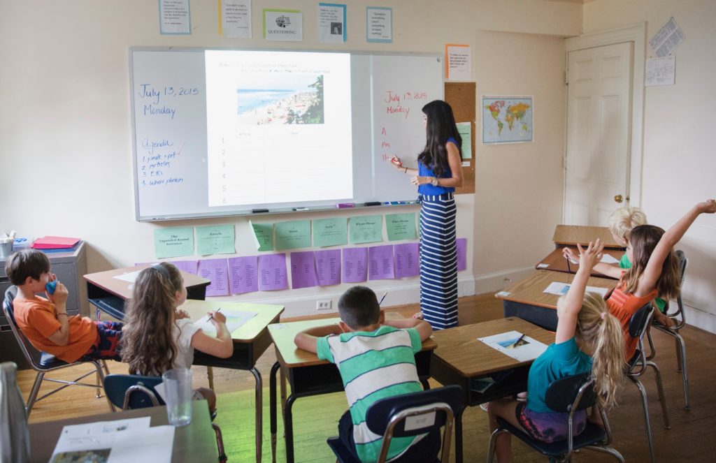 Teacher standing in front of a white board instructing elementary students seated in a horseshoe.