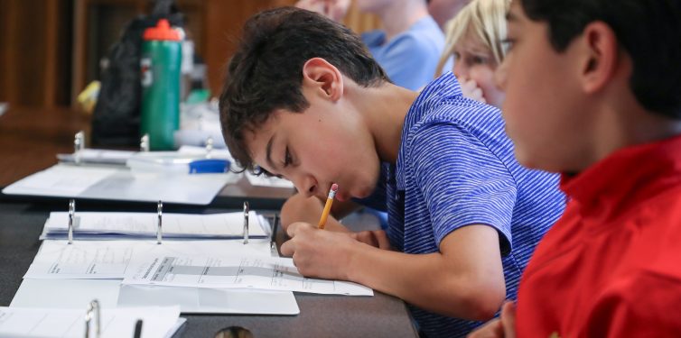 Student leaning over desk taking focused notes with paper and pencil.