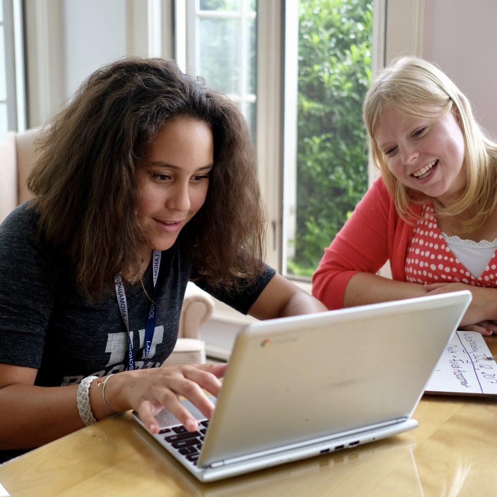 A high school aged student is working one-on-one with a teacher.  The teacher is smiling while peering over the students shoulder to review what is being typed on the student's laptop screen.