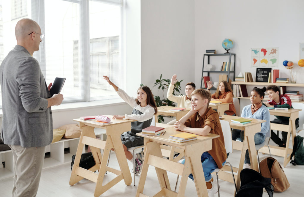 Students seated in rows in a bright classroom have hands raised waiting for the teacher to call upon them.